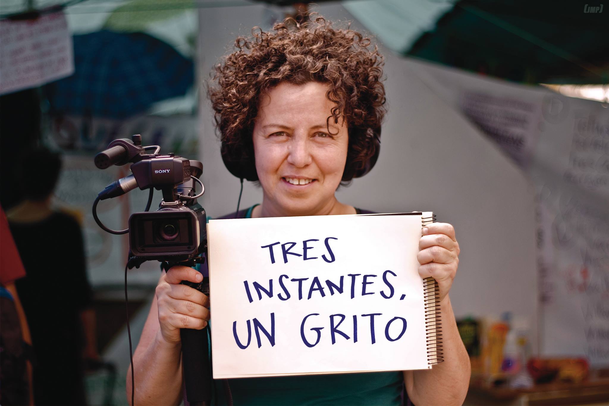 fotografía de una mujer crespa, con audífonos y con un cartel en la mano izquierda que dice "tres instantes, un grito" mientras sostiene una cámara de video con la otra.